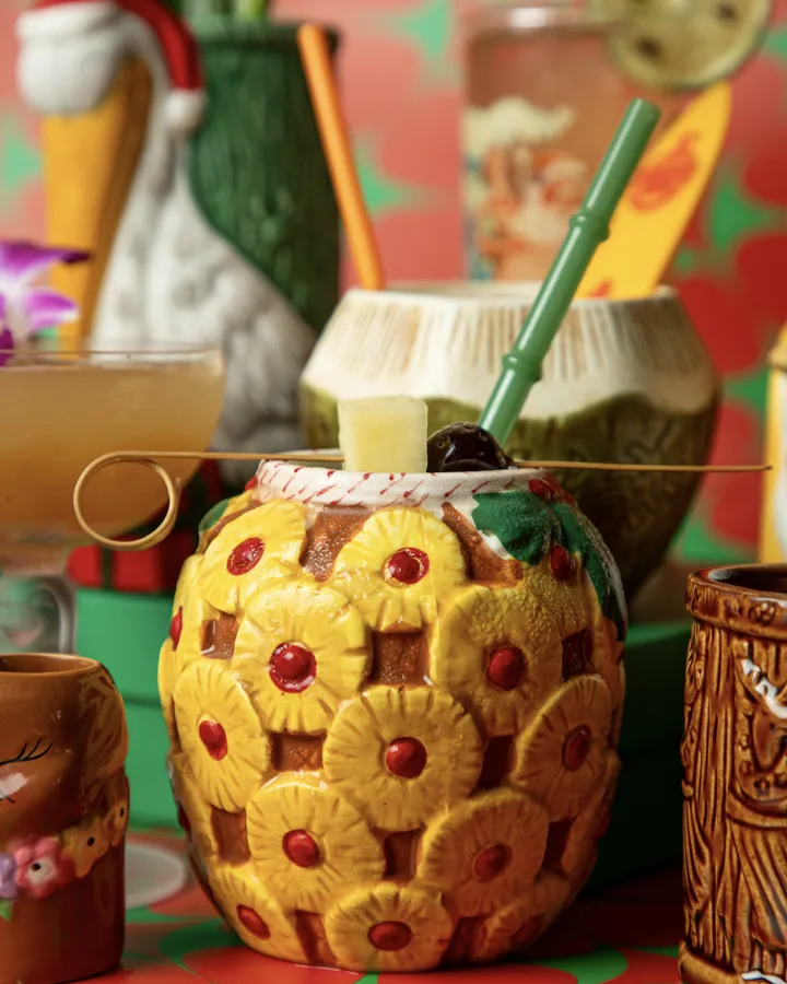 A pineapple tiki mug with a coconut mug and other collection mugs behind it