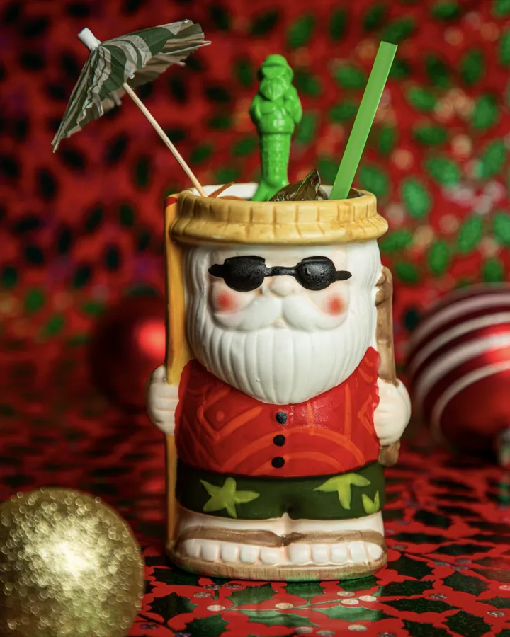 A Santa tiki mug with a cocktail umbrella and green straw on a red holiday backdrop