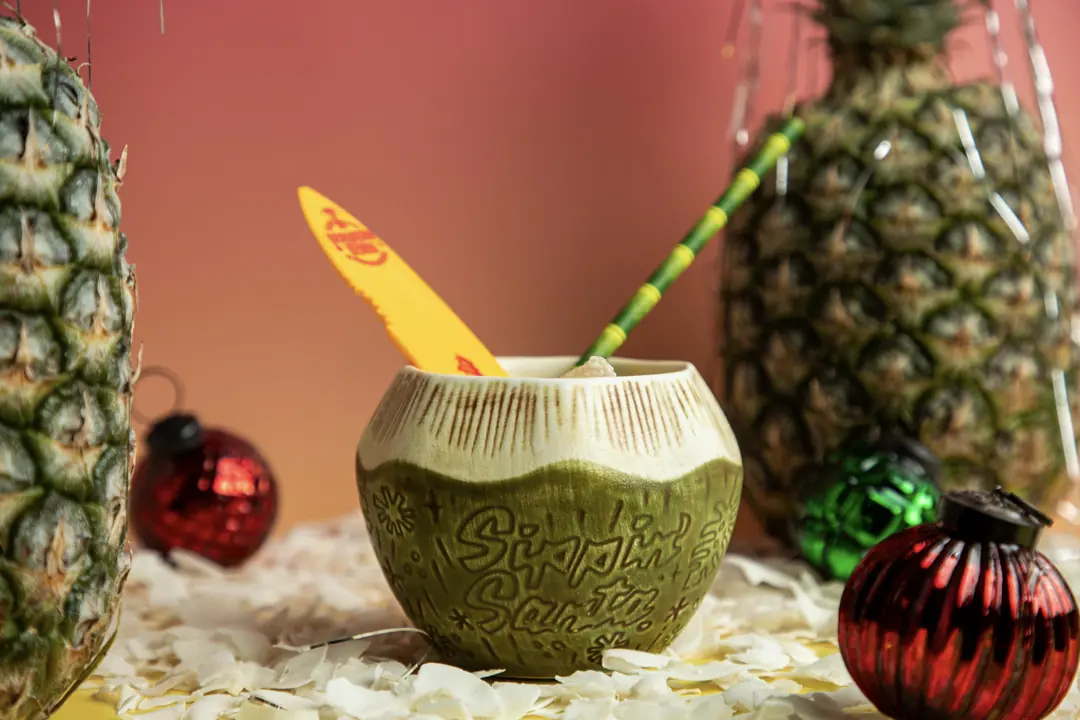 A coconut tiki mug with a yellow surfboard pick, surrounded by pineapples and glass ornaments